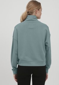Ljusblå mock neck sweatshirt med långa ärmar, ribbad mudd och en avslappnad passform. Slät textur, minimala sömnadsdetaljer på baksidan.