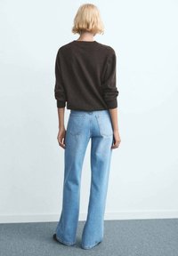 Rjavi pleteni pulover z zavihki na rokavih, kombiniran s svetlo modrimi zvončastimi kavbojkami. Obleka je vidna od zadaj na enostavni podlagi.