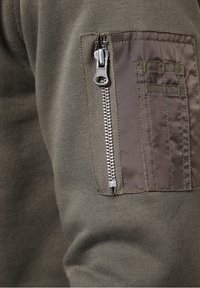 Felpa verde oliva con tessuto testurizzato, tasca con zip sulla manica sinistra e dettagli di cucitura in un colore a contrasto.