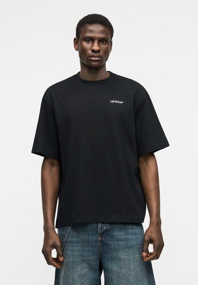 Άνδρας που φοράει μαύρο T-shirt Off-White™ και μπλε τζιν, στέκεται μπροστά σε απλό ανοιχτό γκρι φόντο, κοιτάζοντας προς τα εμπρός με ουδέτερη έκφραση.