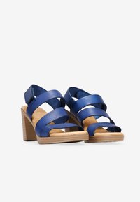 Sandalias de tacón color azul marino con múltiples tiras cruzadas, tacones de bloque con textura de madera y un acabado de cuero suave. Presenta la puntera y la parte trasera al aire.