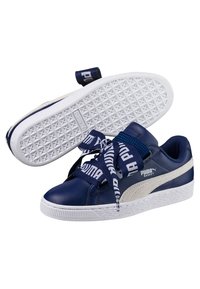 Navy blue Puma Basket sneakers with white soles, beige side stripe, and wide navy laces branded with "Puma" text. One shoe shows textured sole.
