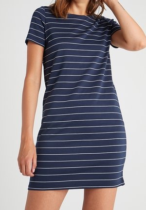 Kobieta ubrana w granatową sukienkę z krótkim rękawem do kolan z cienkimi, białymi, poziomymi paskami, stojąca na jednolitym tle.