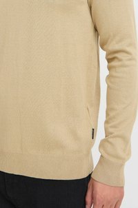 Suéter de punto beige con mangas largas, puños y dobladillo acanalados, que presenta una etiqueta en la costura lateral. Textura suave y diseño ajustado.