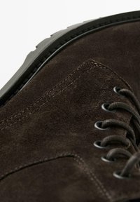 Scarpe in suede marrone con una texture liscia, lacci tondi e suola in gomma. Presentano dettagli di cucitura e occhielli in metallo per i lacci.