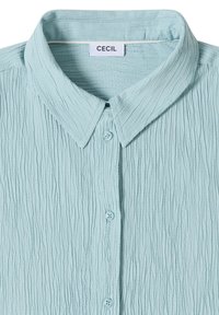 Chemise bleu clair texturée froissée à boutons avec un col pointu et une étiquette « Cecil » à l'intérieur de l'encolure.