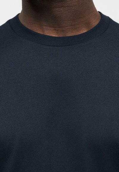 Κοντινή λήψη ενός ατόμου που φοράει μπλούζα σκοτεινού μπλε χρώματος με στρογγυλή λαιμόκοψη, που δείχνει το λαιμό και την άνω περιοχή του στήθους.
