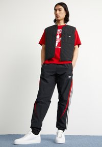 Homem vestindo uma t-shirt vermelha da Adidas, um colete acolchoado preto, calças de treino pretas da Adidas com riscas vermelhas e brancas, e sapatilhas brancas, em pé com as mãos nos bolsos.