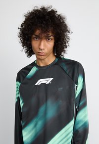 Zwarte longsleeve shirt met teal abstract patroon, voorzien van een witte logo op de borst en contrasterende turquoise accenten langs de naden.