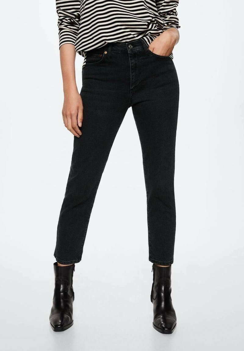 Personne portant un jean skinny taille haute noir, des bottines noires et un haut à manches longues rayé noir et blanc, une main dans la poche.