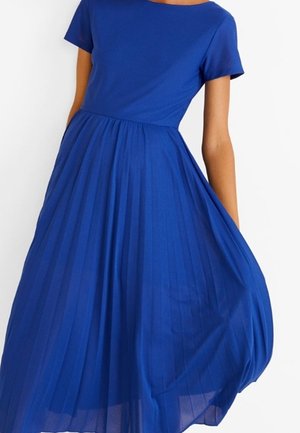 Kobieta ubrana w krótką sukienkę w królewskim odcieniu niebieskiego, z dopasowanym gorsetem i plisowaną, zwiewną spódnicą na białym tle.