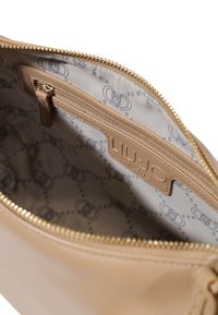 Interno della borsa beige con una cerniera dorata. Presenta una tasca con zip e un'etichetta con logo. Foderato con un tessuto patterned che mostra anelli intrecciati.
