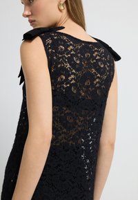 Sort blondekjole med indviklede blomster mønstre, ærmeløs design og silkebue detaljer på skuldrene. Tekstureret og semi-transparent.