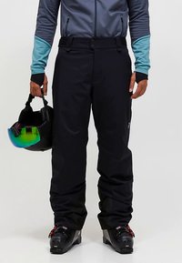 Fekete hó nadrág masszív anyagból, egyenes szabással, két oldalzsebbel és gombos záródással. Sí bakancsokkal és sisakkal párosítva.