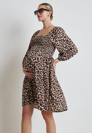 Mujer embarazada que lleva un vestido marrón y beige con estampado de leopardo y gafas de sol, con una mano en su vientre y la otra en su cadera.