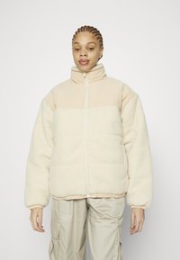 Tossigbeige jacka med hög krage, dragkedjestängning och kontrasterande beige panel högst upp. Har elastiska ärmslut och en mjuk känsla.