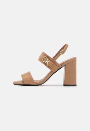 Beżowe sandały na wysokim obcasie z gładką skórzaną cholewką, regulowanym paskiem na pięcie oraz złotym logo CK na przednim pasku. Słupkowy obcas.