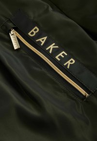 Tela verde oliva con una textura suave, que presenta un zipper dorado y una etiqueta negra que dice "BAKER" en letras doradas.