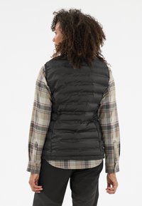 Schwarze, gepolsterte Weste mit horizontalem Steppdesign, getragen über einem karierten Langarmhemd in gedeckten Farben, kombiniert mit dunklen Hosen.