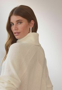 Cremefarvet rullekravesweater med ribbet tekstur og chevronmønster. Modellen bærer sølvfarvede hoops og har bølget brunt hår.