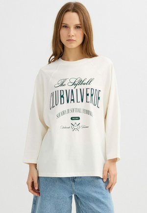 Joven mujer con una camiseta blanca de manga larga con el texto "The Softball Club Valverde" y vaqueros azules, posando frente a un fondo liso.