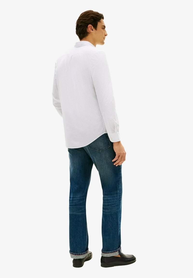Homme portant une chemise blanche texturée à manches longues et un jean bleu, debout, mains détendues, de profil devant un fond clair uni.
