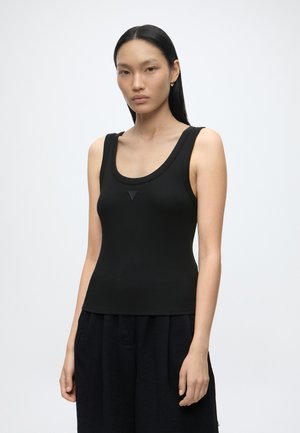 Jeune femme aux longs cheveux raides portant un haut noir sans manches et un pantalon noir, debout devant un fond uni clair.