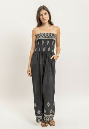 Femme portant une combinaison noire sans bretelles avec des motifs blancs, debout avec une main dans la poche et un bracelet en or au poignet gauche.