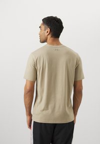 Béžové tričko s krátkym rukávom a okrúhlym výstrihom, s jemnou textúrou a decentným logom potlačeným na zadnej strane. Nosené s čiernymi šortkami.