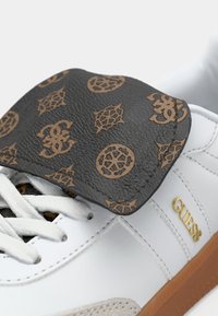 Fehér sportcipő szürke, texturált nyelvvel, arany és barna logómintákkal; arany márkanév díszít és az oldalon.