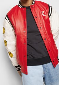 Röd varsityjacka med en glänsande läderkropp, vita ärmar med gula leende ansiktslappar, samt svart- och vitrandig ribbad krage och muddar.