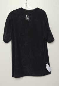 T-shirt noir à manches courtes Nike avec les logos NBA et Nike dans le dos au niveau du col, suspendu à un cintre en plastique transparent sur un fond clair.