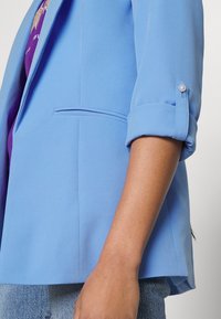 Détail de manche d'une personne portant un blazer bleu clair retroussé sur une chemise violette et un jean bleu, montrant la patte de bouton au poignet.