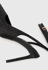 Μαύρες ψηλοτάκουνα παπούτσια με μυτερή μύτη, στολισμένα με ρινοστάτες. Διαθέτουν λεπτό τακούνι στιλέτο και λεπτές λουρίδες γύρω από τον αστράγαλο.