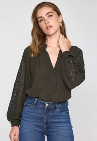Blusa verde scuro con scollo a V, dettagli con bottoni e maniche lunghe con eleganti accenti in pizzo nero. Tessuto morbido e testurizzato.