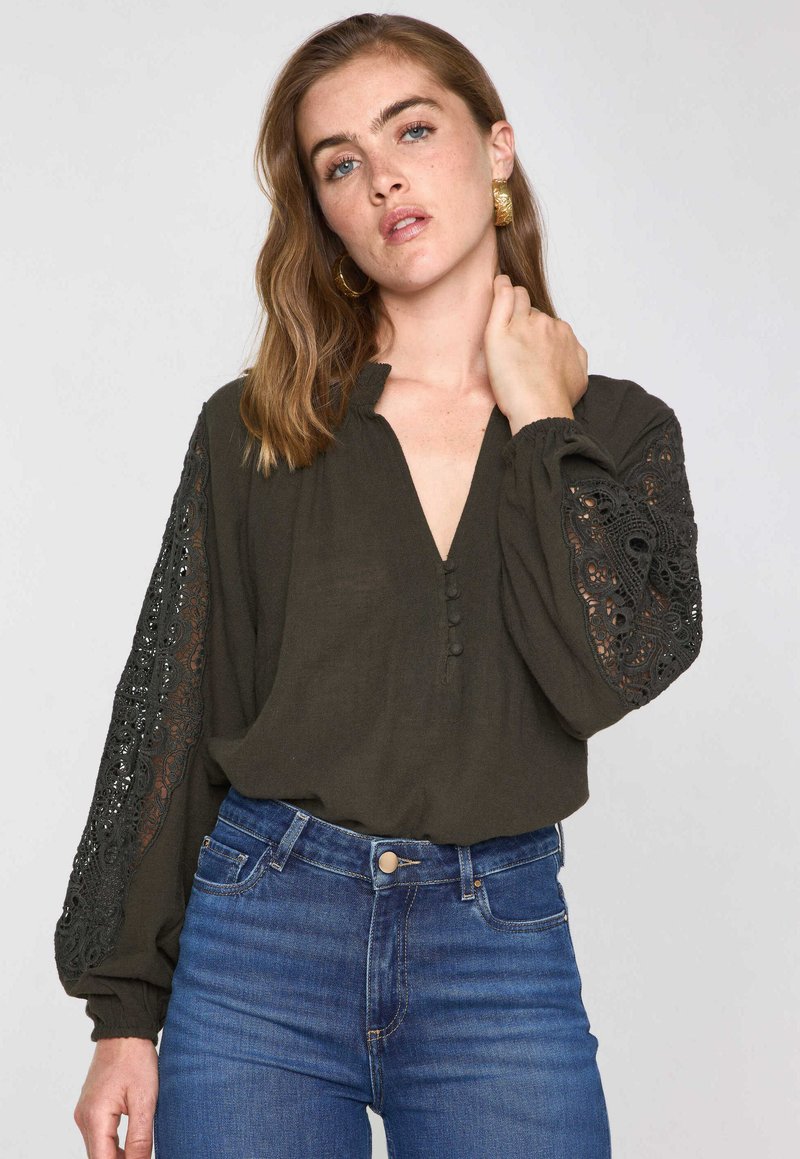 Blusa verde scuro con scollo a V, dettagli con bottoni e maniche lunghe con eleganti accenti in pizzo nero. Tessuto morbido e testurizzato.