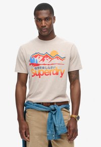 Beige T-shirt med bjerg- og solgrafik i blå, rød og orange, med "Superdry" logo. Bæres med kaki bukser og en denim skjorte bundet om livet.