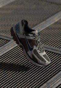 Nike sportcipő, bézs és olívazöld hálós felsőrésszel, fekete Nike logóval és texturált gumitalppal, szürke kiemelésekkel.