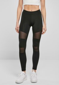 Sorte leggings med gjennomsiktige mesh-paneler ved lårene, glatt stoff, høy midje og en tettsittende design. Kombinert med hvite joggesko.
