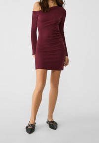 Robe bordeaux décolletée en tissu extensible, design ajusté avec manches longues et détail froncé, associée à des ballerines noires pointues.