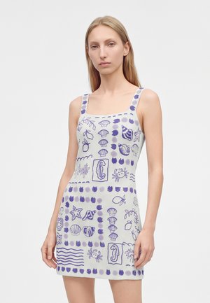Mujer con un vestido blanco sin mangas con patrones azules con temática marina que incluyen conchas, caballitos de mar, cangrejos y estrellas de mar.