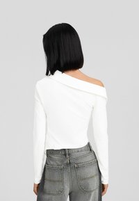 Kvinna med svart bobfrisyr som bär en vit off-shoulder långärmad topp och högmidjade grå jeans, vänd bort från kameran.