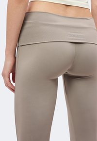 Gros plan d'une personne portant un legging beige avec une taille repliée arborant un petit logo GUESS en strass.