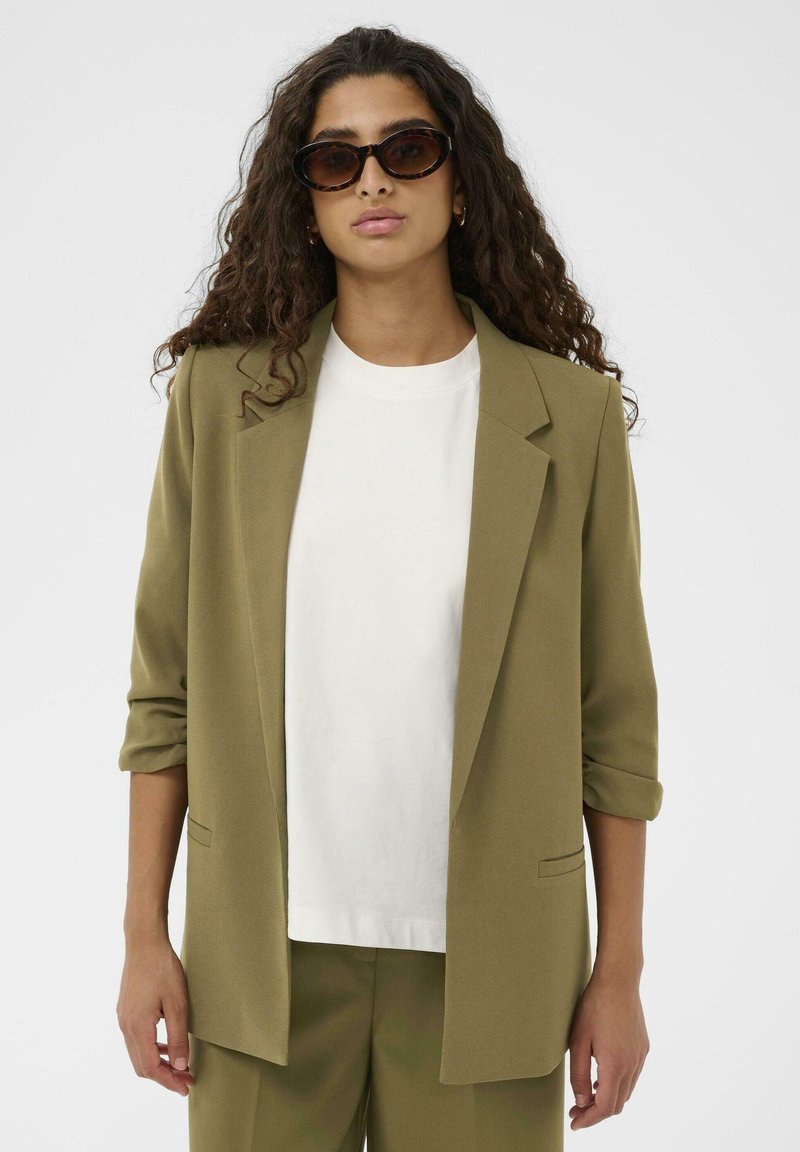 Vrouw met lang, krullend haar draagt schildpadkleurige zonnebril, olijfgroene blazer met opgerolde mouwen, wit T-shirt en bijpassende broek.