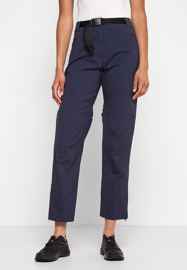 Frau in navyblauen, umwandelbaren Outdoor-Hosen mit verstellbarem Gürtel und schwarzen Sportschuhen, die vor einem weißen Hintergrund steht.