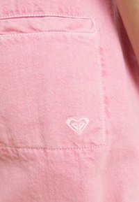 Primer plano de un bolsillo de mezclilla rosa con un logotipo en forma de corazón bordado en blanco en la tela.