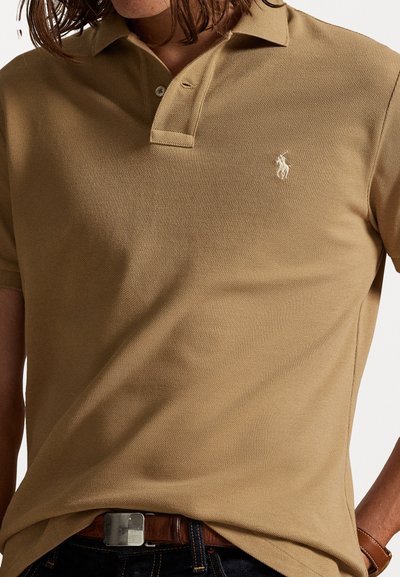 Μπεζ πουκάμισο πόλο από υφασμάτινη υφή, με τριπλό κουμπί και μικρό κεντητό λογότυπο στο αριστερό στήθος.