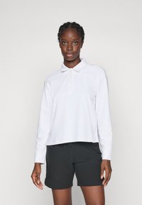 Vrouw met vlechten, gekleed in een witte lange mouwen polo shirt en zwarte shorts, staand tegen een effen lichte achtergrond.