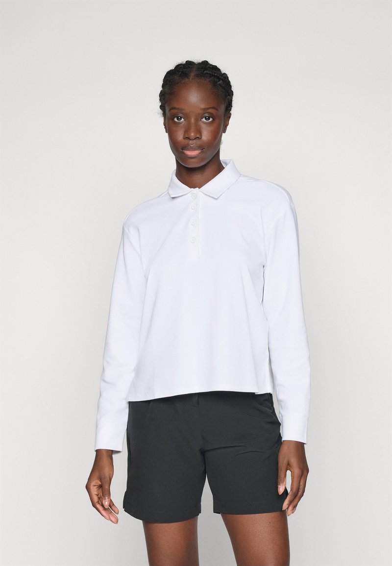 Vrouw met vlechten, gekleed in een witte lange mouwen polo shirt en zwarte shorts, staand tegen een effen lichte achtergrond.