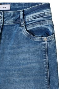 Jean en denim bleu en gros plan montrant la poche avant, les passants de ceinture et les détails de couture sur un tissu texturé.